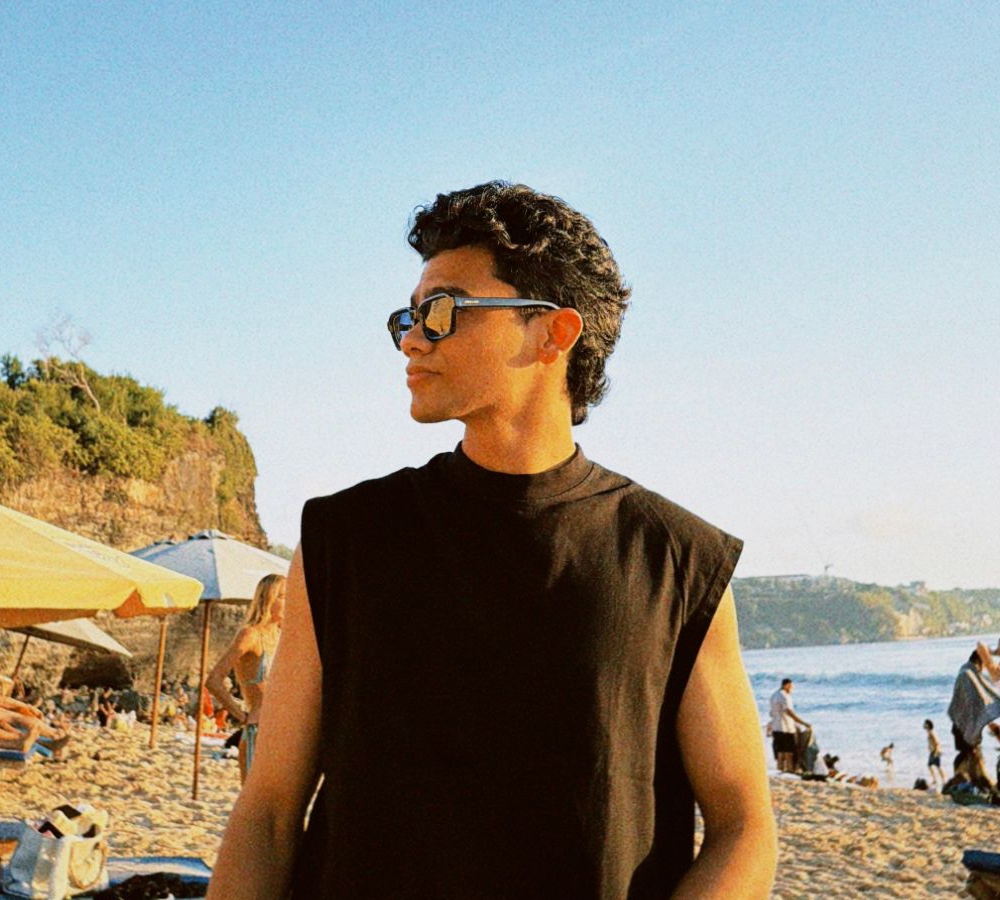 Varun on a beach at golden hour, wearing sunglasses and a black sleeveless tee